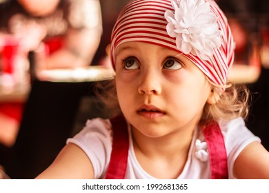 A  3-4 Years Child, Girl With An Expressive Face And Big Eyes Looks Up, Beautiful Little Girl Wearing Dark Red And White Cloths And Flower Head Band, Close Up