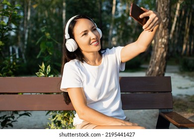 35 Yeaers Old Woman In Headphones Taking Selfie With Her Smartphone While Sitting On The Bench In City Park. Close-up Photo.