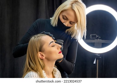 A 35-40 Year Old Female Makeup Artist Wearing A Medical Mask Applies Makeup To A Woman's Face. Concept: Makeup For Women During A Pandemic.