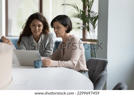 35s and 55s women colleagues working together use computer, meet in office staring at laptop screen, learn new software, younger Hispanic affiliate helps to older with program or app. Tech, business