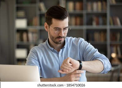 35s businessman wear glasses looks at wristwatch checks time waits for client who is late while sitting at work place desk with laptop. Concept of business, time is money, punctuality and timekeeping - Shutterstock ID 1854710026