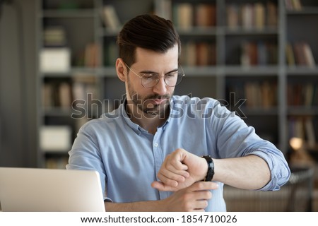 35s businessman wear glasses looks at wristwatch checks time waits for client who is late while sitting at work place desk with laptop. Concept of business, time is money, punctuality and timekeeping