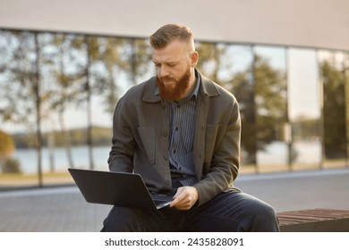 A 35-year-old man with a red beard sits on a bench with a laptop during lunch break. - Powered by Shutterstock