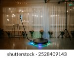 A 360-degree photo booth setup with LED lights and a rotating platform, surrounded by floral arrangements, in front of sheer curtains and string lights.