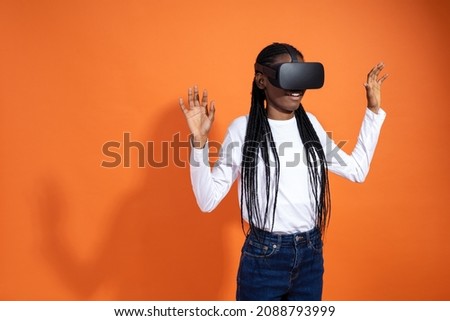 3d technology, virtual reality, entertainment, cyberspace and people concept - frightened african-american girl in vr-glasses seeing something and spreading arms on orange background with copy space