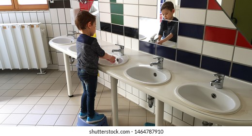3-year-old little boy, dressed in jeans and dark blue longsleeve, washes his hands himself in the sink in the room of the children's toilet, standing in front of the morror. Side view. Selective focus