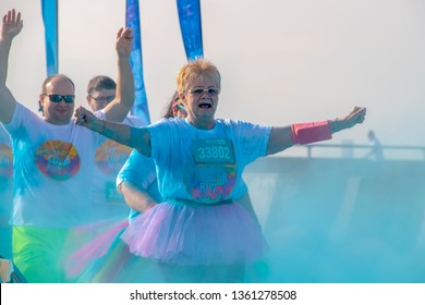 4 - 6 - 2019 - Two Runners On A Senior Citizen Woman In A Tutu And A Balding Man Behind Her - Hold Their Arms Up As They Run Through Colored Powder At A Color Run Race