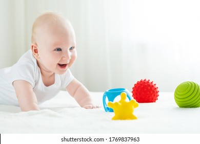 Ein viermonatiger Junge liegt auf dem Bauch, mit Betonung auf den Händen. Er lächelt und spielt mit bunten, lockigen Gummispielzeug.