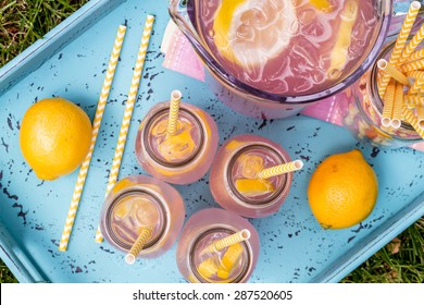 4 Small Glass Bottles And Pitcher Filled With Fresh Squeezed Pink Lemonade With Yellow Swirled Straws And Lemon Slices Sitting On Weather Blue Drink Tray From Above