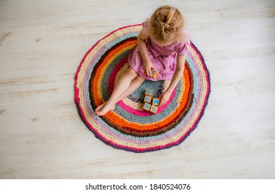 4 Year Old Young Child Sit On Round Colorful Upcycling Crochet Rug From Repurposed T-shirts Yarn On White Laminate Flooring And Play Natural Wood Cubes With Letters. Top Above View.