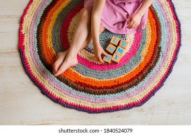 4 Year Old Young Child Sit On Round Colorful Crochet Rug From Repurposed T-shirts Yarn On White Laminate Flooring And Play Natural Wood Cubes With Letters. Top Above View.