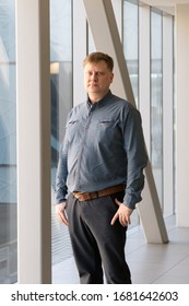 A 40-45-year-old Man In A Blue Plaid Shirt Poses In The Office Back Hallway Amid A Large Window. Portrait Of A Tall Overweight Man Near Large Office Windows.