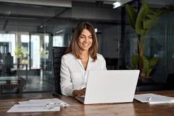 40s Mid Age European Business Woman CEO Using Laptop Application For Work Sitting At Table Workspace In Office. Smiling Latin Hispanic Mature Adult Professional Businesswoman Using Pc Digital Computer