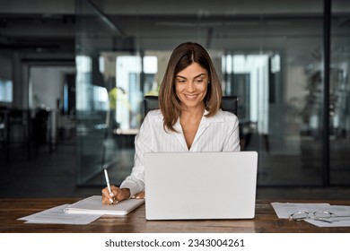 40s mid age European business woman CEO working on laptop sitting at table workspace, making notes in office. Smiling Latin Hispanic mature adult professional businesswoman using pc digital computer