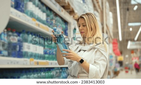 40s woman buy still water bottle. Girl take aqua item put cart. Health care row. Person go detox shop. Lady drink liter daily. Customer walk food store. Buyer pick up clean water. Many plastic bottles
