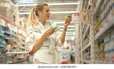 40s woman buying baby food. Mum read fruit mash composition. Girl look meal ingredients. Mother choose kid jar shop. Lady check foods exp date. Baby store row. Parent take puree put cart. Pick up item - Powered by Shutterstock