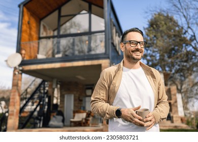 A 40-year-old adult man stands confidently in front of his modern home holding a cup of tea or coffee smiling He looks content and happy with his life and proud of his house copy space - Powered by Shutterstock