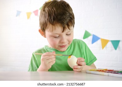 A 4-5 Year Old Boy Sits At A Table And Paints A Wooden Easter Egg With Yellow Paint.The Child Is Absorbed In Business, Smiling And Not Looking At The Camera.Cute Child Is Busy With Creativity,drawing