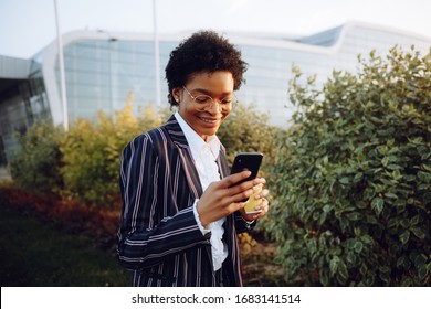 4k. Travel, Digital. Charming African American Woman In Elegant Striped Suit, Holding Phone And Cup Of Coffee. Stands On Background The Bushes Near Airport. Businesswoman Or Model. And Looks In Phone