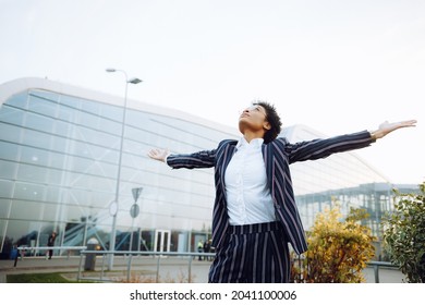 4k. Travel, Digital. A charming African American woman in an elegant striped suit, looks at the sky, smiles and feels fantastic, her arms are wide. It stands against the bushes near the airport