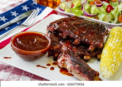 4th Of July Celebration Picnic With Barbeque Pork Ribs With Sauce Sitting On White Plate With Ear Of Corn And Side Serving Of Salad