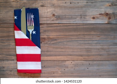 4th Of July Rustic Picnic Table Place Setting With Silverware And American Flag Napkin With Background Of Wood Boards For Copy Space.  