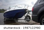 A 4x4 black car pulls blue-white trimaran cabin motor boat on trailer on concrete slipway into the river water, boat launch before active recreation on the river