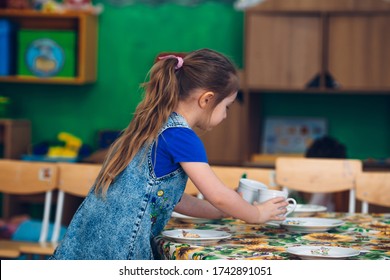 A 4-year-old Girl With Blond Hair Of European Appearance Helps To Set The Table On Duty In The Kitchen Puts Cups, Spoons, Cutlery. Regime Moments In Kindergarten. Child Education.