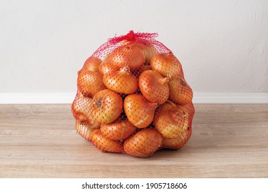 5 Kg Farm Onions In A Red Pp Mesh Bag. Polypropylene Net Sack With 11 Lb Of Organic Onions On A Brown Floor Indoors. Buying Fresh Vegetables In Bulk. Front View.