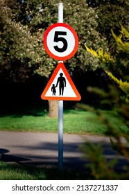 A 5 Mile Per Hour Round Road Sign Above A Parent And Child Triangle Sign Against A Blurry Park Background