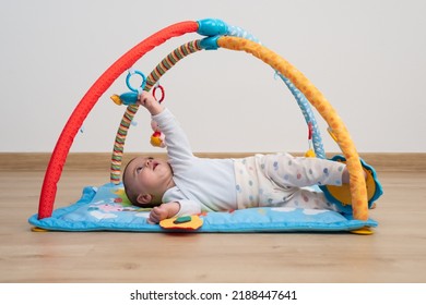 5 Months Baby Lying Face Up On A Blue Play Mat While Trying To Catch A Toy From A Baby Gym. Montessori Activities.