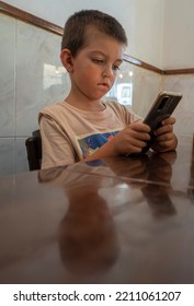 5 Year Old Boy Concentrating His Attention On His Mobile Phone, Holding His Mobile Phone With Both Hands And Pressing His Mouth. He Is Sitting At A Wooden Table With A Wall Of White Tiles Behind Him.