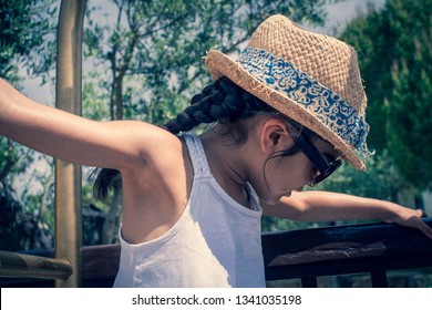 A 5 Year Old Girl In A Sunhat And Sunglasses Plays On The Luggage Rack Of A Bellboy, Outside A Holiday Hotel, Whilst On Vacation. 