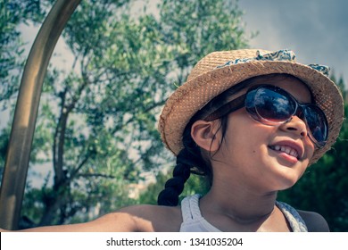 A 5 Year Old Girl In A Sunhat And Sunglasses Plays On The Luggage Rack Of A Bellboy, Outside A Holiday Hotel, Whilst On Vacation. 