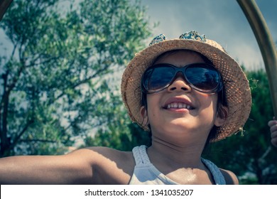 A 5 Year Old Girl In A Sunhat And Sunglasses Plays On The Luggage Rack Of A Bellboy, Outside A Holiday Hotel, Whilst On Vacation. 