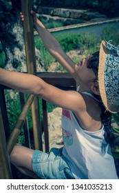 A 5 Year Old Girl In A Sunhat And Sunglasses Plays On The Luggage Rack Of A Bellboy, Outside A Holiday Hotel, Whilst On Vacation. 