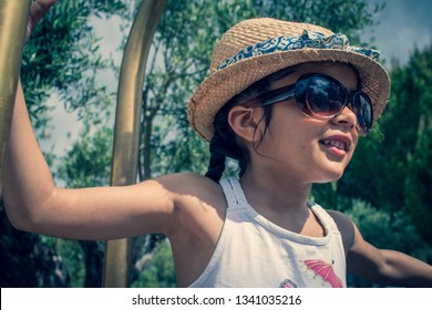 A 5 Year Old Girl In A Sunhat And Sunglasses Plays On The Luggage Rack Of A Bellboy, Outside A Holiday Hotel, Whilst On Vacation. 