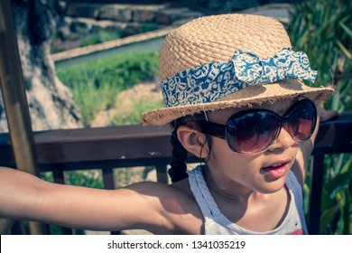 A 5 Year Old Girl In A Sunhat And Sunglasses Plays On The Luggage Rack Of A Bellboy, Outside A Holiday Hotel, Whilst On Vacation. 