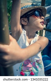 A 5 Year Old Girl In A Sunhat And Sunglasses Plays On The Luggage Rack Of A Bellboy, Outside A Holiday Hotel, Whilst On Vacation. 