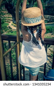 A 5 Year Old Girl In A Sunhat And Sunglasses Plays On The Luggage Rack Of A Bellboy, Outside A Holiday Hotel, Whilst On Vacation. 