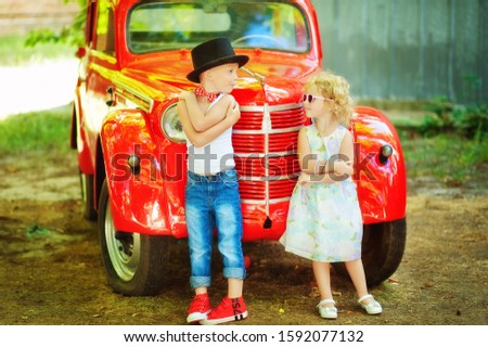 5 years old boy in blue jeans, white t-shirt, red bow tie and black cylinder and 3 years old girl in light dress stay near red retromobile old car and look at each other. Happy children outdoor.