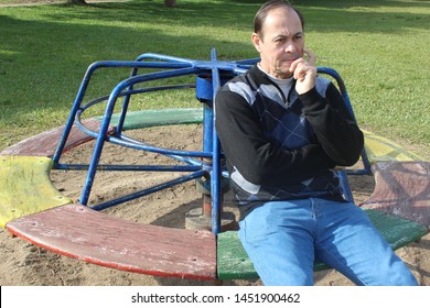 50 Year Old Man Sitting On A Spinning Tour In A Playground Of A Beautiful Square, Colorful Toy In Grass, Sad And Worried Physiognomy