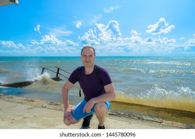 50 Year Old Man Squatting On The Background Of The Raging Sea And Waves, In Good Sunny Weather.