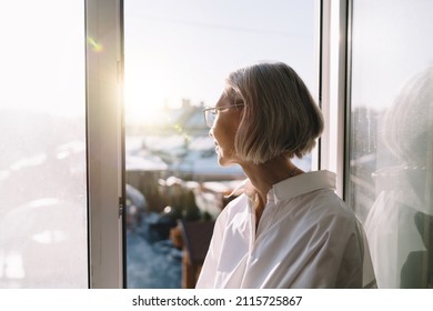 50 Years Old Caucasian Woman Looking From Window On Blur City. Concept Of Domestic Lifestyle. Modern Successful Woman. Cropped Image Of Grey Hair Lady Wearing Shirt And Glasses. Sunny Day