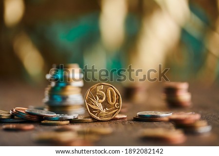 A 50 ZAR cents coin standing up on a brown wood surface and gold and blue background with other currencies coins surrounding and towering up in stacks.