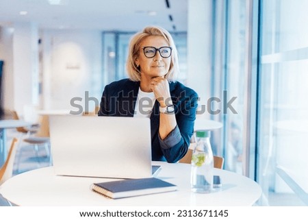 50's confident mature businesswoman dreamy looking at window, middle-aged experienced senior female professional working on laptop in open space office. Female entrepreneur working remotely.