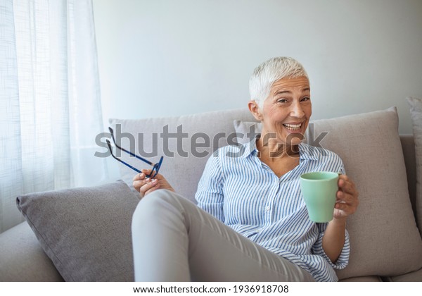 50-year-old woman at\
home drinking tea. Cosy looking senior woman at home with hot\
drink. Happy vivacious middle-aged blond woman holding a cup of tea\
or coffee looking at the\
camera