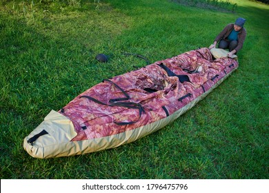 55-60 Year Old Man Lays Out An Inflatable Kayak On The Bright Green Grass. The Boat Is Narrow And Long. Clear Sunny Day, Outdoors, Preparing For A Sailing Trip