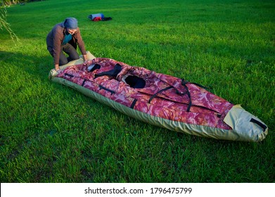 55-60 Year Old Man Lays Out An Inflatable Kayak On The Bright Green Grass. The Boat Is Narrow And Long. Clear Sunny Day, Outdoors, Preparing For A Sailing Trip