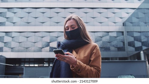 5G internet connection. Young beautiful business woman in face mask using smart phone app at large futuristic building.